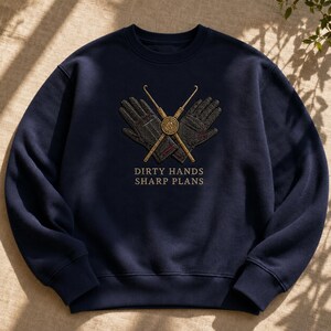 May include: A navy blue crewneck sweatshirt featuring an embroidered graphic of two dark gloves crossed behind two gold-colored crochet hooks. Below the graphic, the text reads "DIRTY HANDS SHARP PLANS" in gold-colored capital letters.