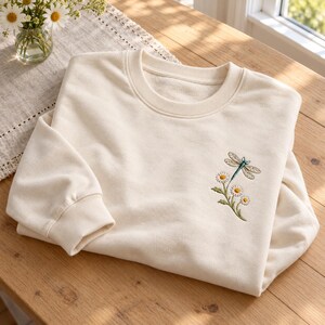 May include: A cream-colored crewneck sweatshirt featuring an embroidered design on the chest. The embroidery depicts a blue and green dragonfly hovering above a small cluster of white daisies with yellow centers and green stems and leaves.