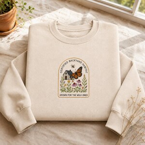 May include: A beige crewneck sweatshirt featuring an embroidered arch-shaped design. The graphic depicts a small house, a monarch butterfly, a bee, and various wildflowers. The text reads "CERTIFIED BACKYARD HABITAT" and "GROWN FOR THE WILD ONES."