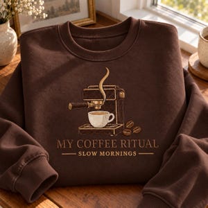 May include: A dark brown crewneck sweatshirt featuring an embroidered graphic of an espresso machine with a steaming cup of coffee and three coffee beans. The text reads MY COFFEE RITUAL SLOW MORNINGS in a clean, sans-serif font below the design.
