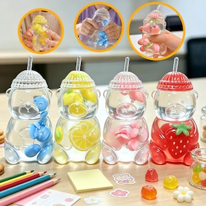 May include: Four clear bear-shaped plastic water bottles with straws and lids. Each bottle contains floating fruit-themed decorations: blue berries, yellow lemon slices, pink flowers, and a red strawberry. The bottles are arranged in a row.
