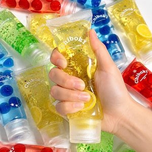 Peut inclure: Une collection de tubes souples transparents remplis de gel coloré, de tranches de fruits et de paillettes en forme d'étoile. Chaque tube porte le texte Chiboky et un bouchon à vis. Les gels sont disponibles en jaune, bleu, vert et rouge.