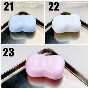 May include: Three smooth, rectangular bars of soap with rounded edges and a central indentation. Item 21 is light blue, item 22 is white, and item 23 is light pink. Each bar is displayed individually on a metallic tray with a reflective surface.
