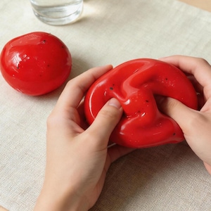 May include: Two bright red, glossy, stress-relieving toys. One is round, the other is being squeezed by hands. The toys have small black speckles. A glass of water is in the background. The toys are on a light beige surface.