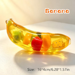 May include: A clear yellow banana-shaped resin object containing embedded colorful fruit pieces including green kiwi, orange, red, and yellow segments. The text "Banana" appears above, and the text "Size: 16*4cm/6.28*1.57in" is below.