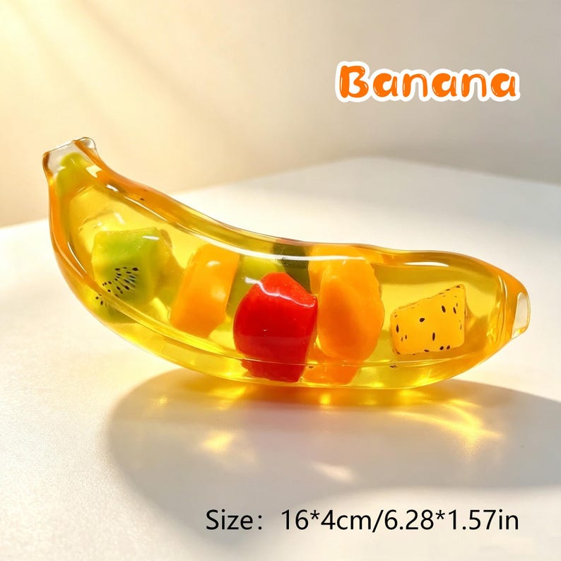 May include: A clear yellow banana-shaped resin object containing embedded colorful fruit pieces including green kiwi, orange, red, and yellow segments. The text "Banana" appears above, and the text "Size: 16*4cm/6.28*1.57in" is below.