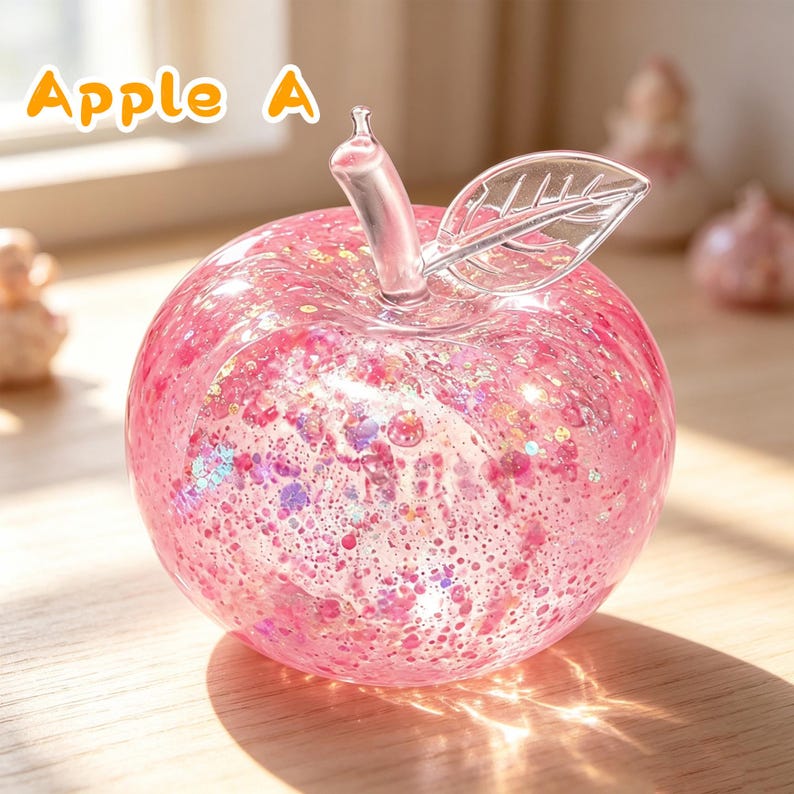 May include: A clear glass apple figurine filled with pink liquid, shimmering glitter, and small pink particles. The apple features a transparent glass stem and a single clear glass leaf attached to the top. The text Apple A is visible.