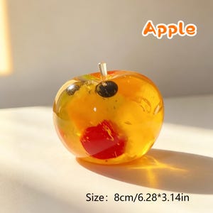 May include: A translucent, amber-colored apple-shaped object with embedded colorful fruit pieces, including a prominent red segment and dark spots. The text 'Apple' appears above, and the text 'Size: 8cm/6.28*3.14in' is printed below the object.