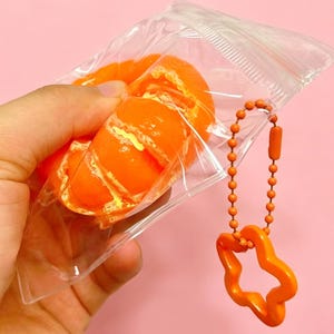 Op de afbeelding: Oranjegekleurde, squishy speelgoed mandarijn sinaasappelschijfjes in een doorzichtige plastic zak met ritssluiting. Een oranje sleutelhanger met een stervormige bedel hangt aan de zak. De achtergrond is effen roze.