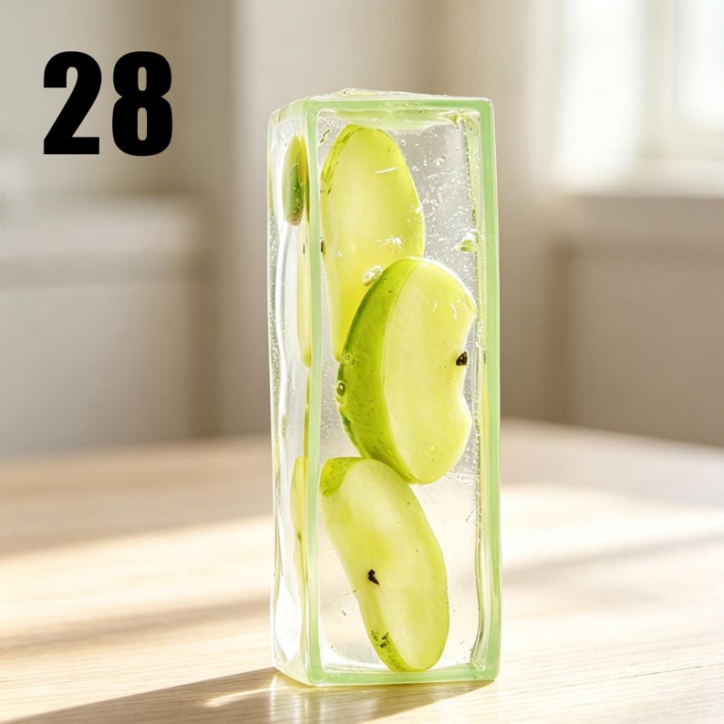 May include: A rectangular clear resin block containing several sliced green apple pieces suspended within the material. The number 28 is printed in black in the upper left corner. The object has a smooth, transparent finish with visible air bubbles.
