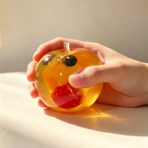 May include: A round, translucent amber-colored glass apple paperweight featuring a clear glass stem at the top. Inside the glass, there are embedded dark circular shapes and a bright red spherical element visible through the clear material.