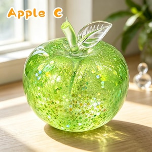 May include: A clear glass apple sculpture featuring a vibrant green speckled interior with embedded glitter particles. The apple has a smooth, rounded body, a translucent green stem, and a single clear glass leaf with etched vein details. The text Apple C is visible.