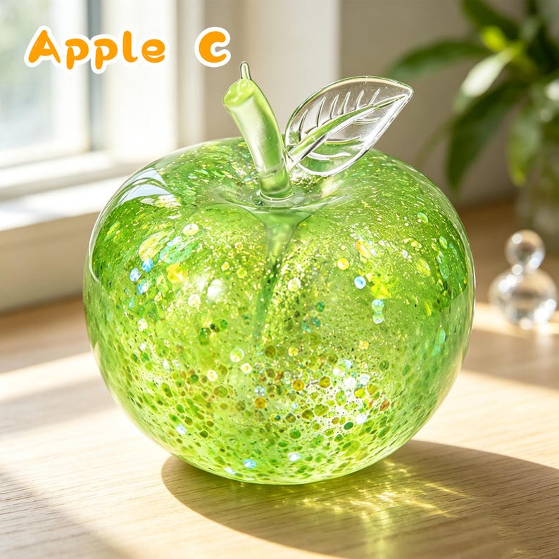 May include: A clear glass apple sculpture featuring a vibrant green speckled interior with embedded glitter particles. The apple has a smooth, rounded body, a translucent green stem, and a single clear glass leaf with etched vein details. The text Apple C is visible.