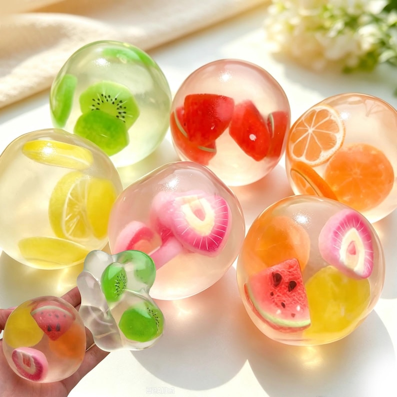 May include: A collection of clear, spherical stress balls filled with various fruit-shaped inserts, including slices of kiwi, watermelon, lemon, orange, and strawberry. The translucent spheres vary in size, with some held in a hand for scale.