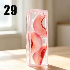 May include: A rectangular clear glass vase with a light pink frame containing three sliced peach segments suspended in a transparent gel-like substance. The number 29 is printed in black in the upper left corner of the image.