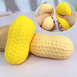 Puede incluir: Dos bolas antiestrés con forma de cacahuete, amarillas y beige. La amarilla está en posición vertical, mientras que la beige está en horizontal. Una imagen más pequeña muestra una mano apretando una bola amarilla y otra sujetando una beige.