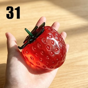 May include: A glass strawberry figurine featuring a translucent red body with a textured surface of small indentations and a dark green glass stem at the top. The number 31 is printed in black in the upper left corner of the image.