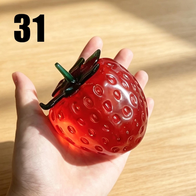 May include: A glass strawberry figurine featuring a translucent red body with a textured surface of small indentations and a dark green glass stem at the top. The number 31 is printed in black in the upper left corner of the image.