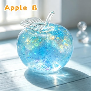 May include: A clear glass apple figurine filled with blue and iridescent glitter particles, featuring a smooth glass stem and a single leaf detail. The text "Apple B" is printed in orange in the upper left corner of the image.