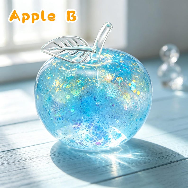 May include: A clear glass apple figurine filled with blue and iridescent glitter particles, featuring a smooth glass stem and a single leaf detail. The text "Apple B" is printed in orange in the upper left corner of the image.