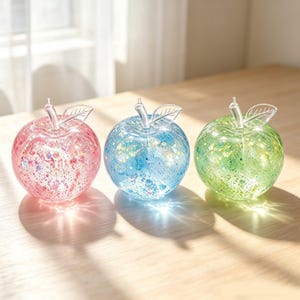 May include: Three round glass apple figurines in pink, blue, and green. Each apple features a clear glass stem with a single leaf and is filled with internal glitter and confetti particles that catch the light, creating a sparkling effect.