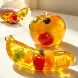 Può includere: Un piatto in resina gialla trasparente a forma di banana e un fermacarte a forma di mela in resina gialla trasparente, entrambi contenenti pezzi di frutta colorati incastonati come kiwi, arancia e segmenti rossi, con un piccolo stelo sulla mela.