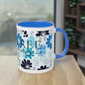 Könnte beinhalten: Eine weiße Keramiktasse mit blauem Rand und Henkel. Die Tasse zeigt ein blaues und weißes Blumenmuster mit dem Text "FREE" in schwarzen Buchstaben.