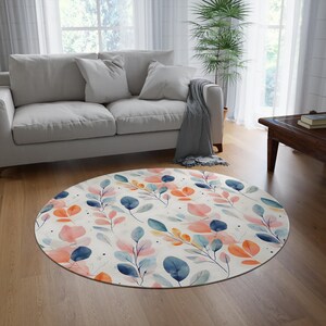Könnte beinhalten: Ein runder, weißer Teppich mit einem Aquarellmuster aus blauen, rosa und orangen Blättern. Der Teppich liegt auf einem Holzboden in einem Wohnzimmer.