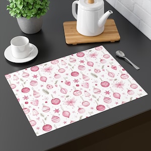 Könnte beinhalten: Ein weißer Tischset mit einem sich wiederholenden Muster aus rosa Weihnachtskugeln, Sternen und floralen Elementen. Das Design umfasst verschiedene Kugelformen, Schleifen und Zweige auf weißem Hintergrund. Eine Tasse und Untertasse, eine Teekanne und ein Löffel sind ebenfalls sichtbar.