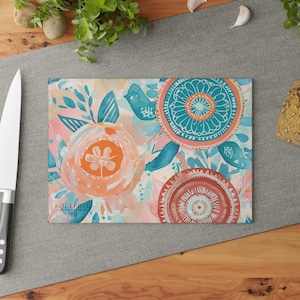 Könnte beinhalten: Ein Schneidebrett aus Glas mit einem farbenfrohen Blumendesign. Das Design zeigt türkisfarbene, orangefarbene und rosafarbene Aquarellblumen und Kreise. Der Text "SOULFREE" ist auf das Brett gedruckt.