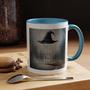 Könnte beinhalten: Eine weiße Keramiktasse mit blauem Rand und Henkel. Die Tasse zeigt einen schwarzen Hexenhut mit einem Besen und dem Text "YES I CAN DRIVE A STICK".