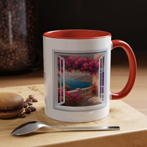 Könnte beinhalten: Weiße Keramik-Kaffeetasse mit rotem Rand und Henkel. Die Tasse zeigt ein farbenfrohes Bild eines Fensters mit Blick auf einen Balkon, der auf ein blaues Meer blickt. Rosa Blumen hängen vom Balkon.