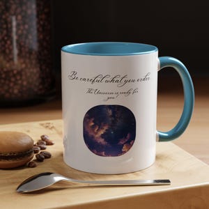Könnte beinhalten: Weiße Keramiktasse mit tealfarbenem Rand und Henkel. Die Tasse zeigt ein Zitat, das lautet: "Sei vorsichtig, was du bestellst. Das Universum ist bereit für dich!" und ein Foto eines sternenklaren Nachthimmels.