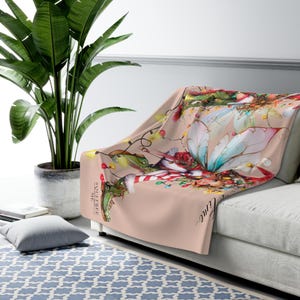 Pode incluir: Uma manta de arremesso rosa com um design de fada caprichoso que apresenta uma fada com asas, uma bota verde e uma meia vermelha e branca listrada. A manta tem um tema natalino festivo com luzes e o texto "É a época mais maravilhosa..." impresso nela.