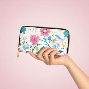 Könnte beinhalten: Eine weiße Reißverschluss-Geldbörse mit einem Blumendruck mit rosa, blauen und grünen Aquarellblumen. Die Geldbörse hat einen goldenen Reißverschluss.