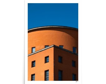 May include: Architectural print featuring a vibrant orange building against a clear blue sky. The building has a rounded top and a rectangular lower section with multiple windows. The image is a vertical composition.