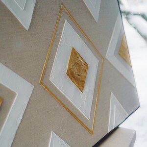 Könnte beinhalten: Abstrakte geometrische Leinwandkunst mit einem neutralen beigefarbenen Hintergrund und weißen und goldenen Rautenformen. Die Rauten sind goldfarben umrandet und haben eine strukturierte Oberfläche, die ein modernes, minimalistisches Design erzeugt.