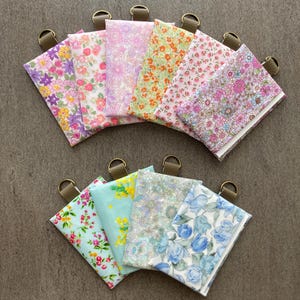 以下が含まれることがあります： ピンク、紫、オレンジ、青など、さまざまな色の花柄生地のポーチのコレクション。各ポーチには、茶色のストラップに小さな金色のリングが付いています。