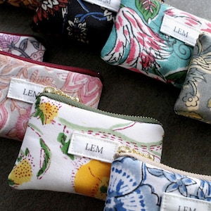 Puede incluir: Una colección de pequeñas bolsas rectangulares con cremallera y varios diseños florales y estampados. Cada bolsa tiene una cremallera dorada y una pequeña etiqueta blanca con la palabra "LEM". Las bolsas son en tonos de rosa, azul, verde y amarillo.