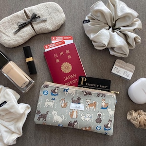 May include: A gray zippered pouch with a white and brown dog pattern. The pouch is open and has a Japanese passport, a Priority Pass card, and a LEM tag inside. The pouch is on a gray surface with other travel items, including a red passport, a pair of eye masks, a hair scrunchie, and a small white case.