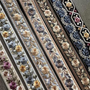 May include: Five strips of decorative fabric trim featuring intricate floral embroidery. The designs include various color palettes such as light blue, beige, grey, and black, accented with metallic thread and sequins in a repeating botanical pattern.
