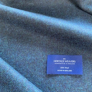 May include: Close-up of a textured teal and navy blue fabric with a herringbone pattern. A blue label reads "HERITAGE WEAVERS HUDDERSFIELD + ENGLAND 100% Wool WOVEN IN ENGLAND". The fabric is likely wool and suitable for apparel or home decor.