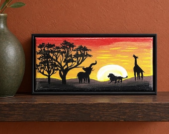 May include: A rectangular canvas painting in a black frame depicting a savanna landscape at sunset. The scene features black silhouettes of trees, an elephant, a lion, and a giraffe against a vibrant red, orange, and yellow sky with a large sun.