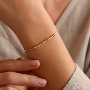 May include: A delicate gold bracelet featuring a series of small, cylindrical and spherical beads. The bracelet is worn on a wrist, with the skin tone appearing fair. The jewelry is simple and elegant, suitable for everyday wear.