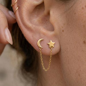 May include: Gold-colored celestial earrings featuring a crescent moon and a star, connected by a delicate chain. The earrings are worn on an ear, showcasing the jewelry's design and detail. The earrings are a stylish accessory.