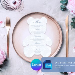 Könnte beinhalten: Eine weiße, bärenförmige Menükarte mit dezentem Blumenmuster und blauem Text, der Pauline's Baby Shower July 27, 2022 lautet, platziert auf einem runden Teller mit goldener Gabel und Messer, umgeben von rosa Blumen auf einem hellgrauen Untergrund.