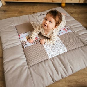 Könnte beinhalten: Eine Patchwork-Spielmatte in Beige- und Brauntönen mit einem Baby darauf. Die Matte hat ein gestepptes Design mit floralen und strukturierten Stoffquadraten. Der Name "Maleo" ist auf die Matte gestickt. Das Baby trägt einen gestreiften Strampler.