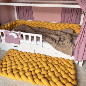 Op de afbeelding: Een wit houten bedframe met een mosterdgele gevlochten bumper en bijpassend vloerkleed. Het bed heeft een bruin deken en een roze gordijn. Een roze zakorganizer hangt aan de zijkant van het bed.