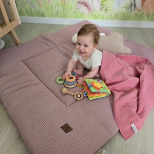 May include: A baby smiles while playing on a mauve waffle-textured play mat. The mat has a quilted design and a small brown label. Toys include a wooden rattle, a colorful soft book, and a ring stacker. A pink blanket is draped nearby.