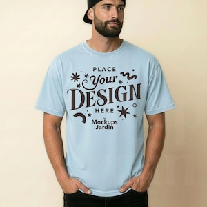 May include: A light blue t-shirt with the text "PLACE Your DESIGN HERE Mockups Jardin" in dark brown. The shirt has short sleeves and is being worn with black jeans and a black baseball cap.